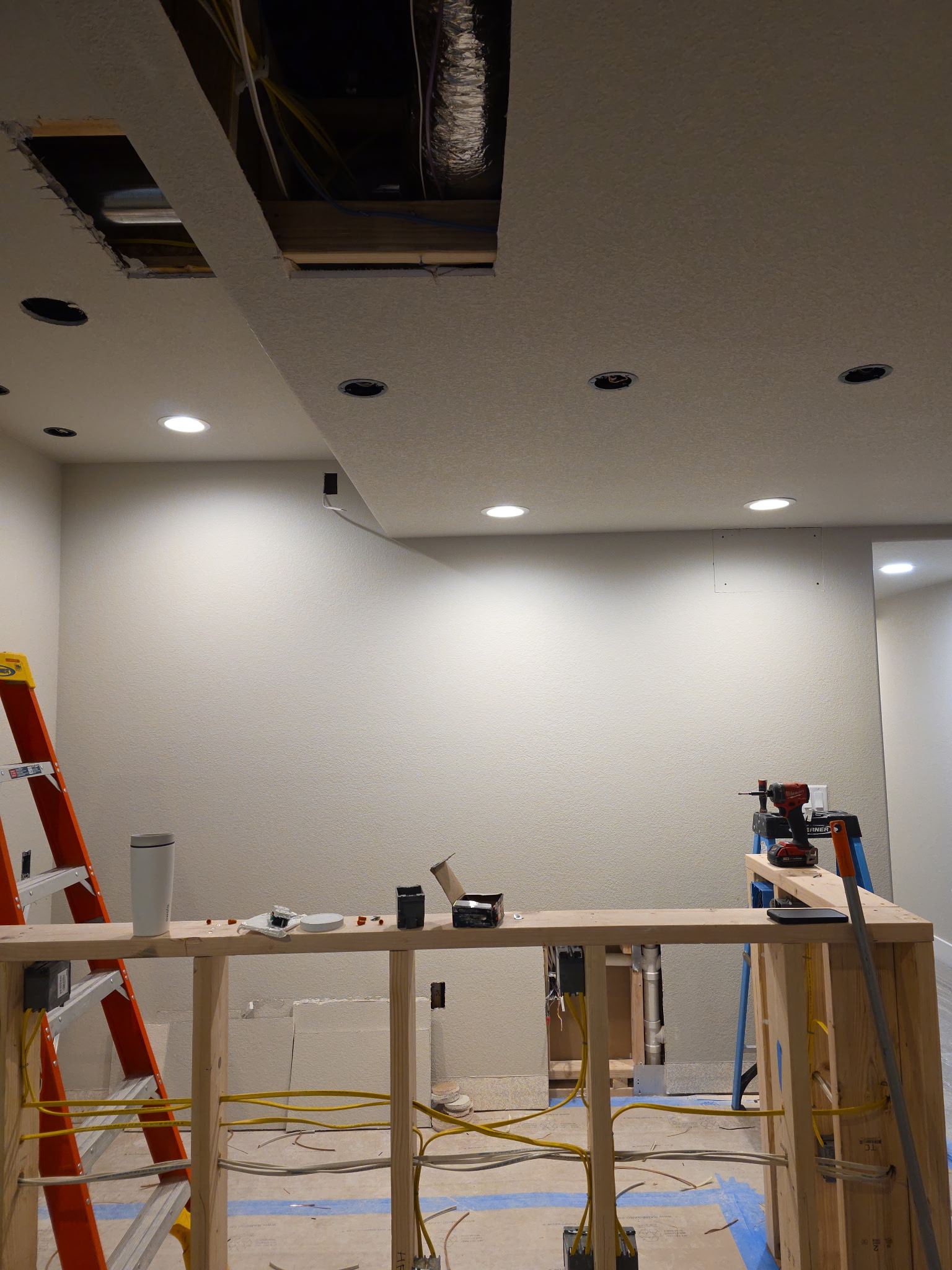 Room under construction with exposed ceiling wiring, recessed lights, a red ladder, and tools on a partially built wooden counter.