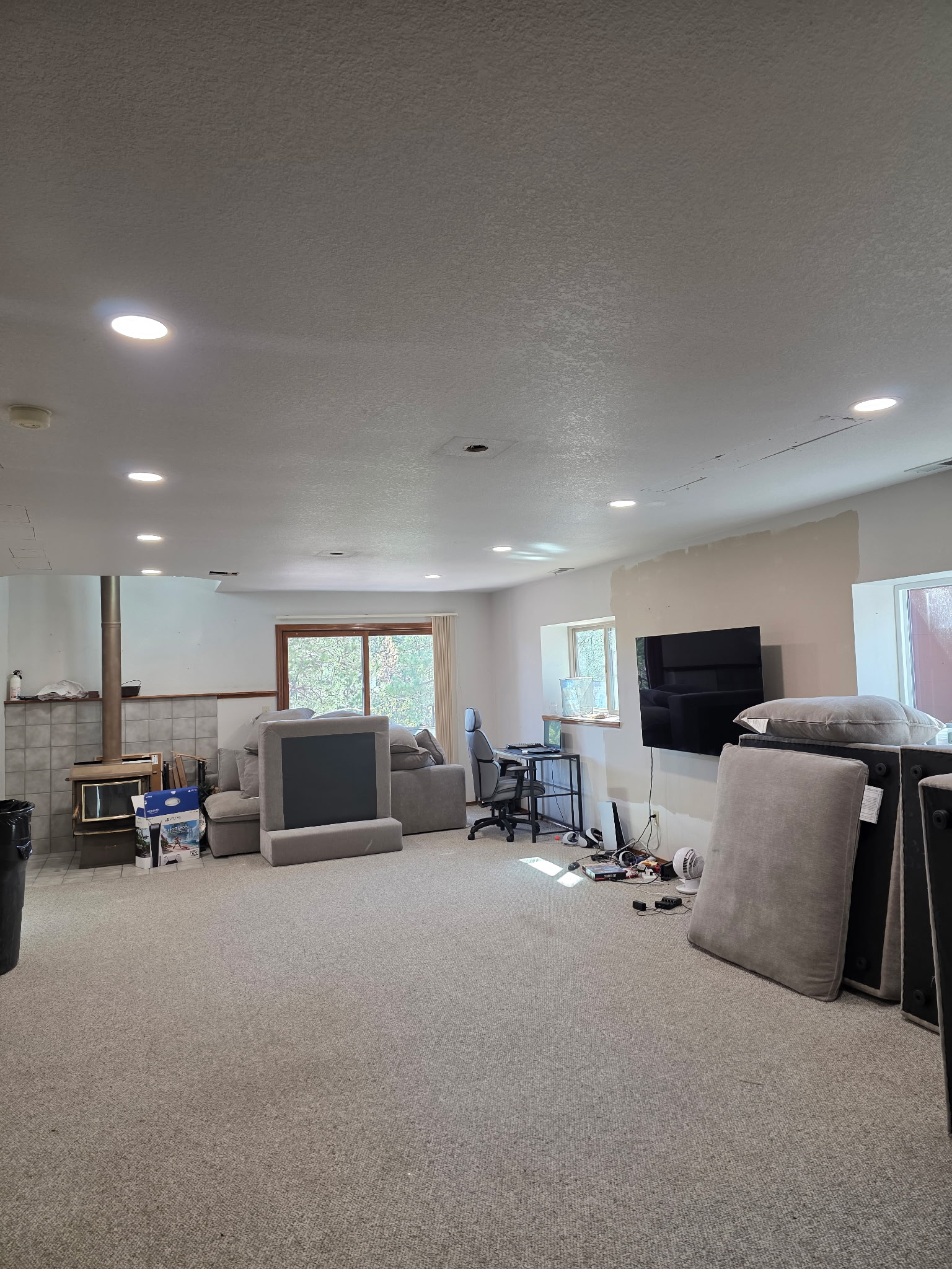 A spacious, carpeted room with recessed LED lighting installation, a wood stove, mounted TV, scattered furniture cushions, a desk with a chair, and boxes near large windows and a sliding glass door.