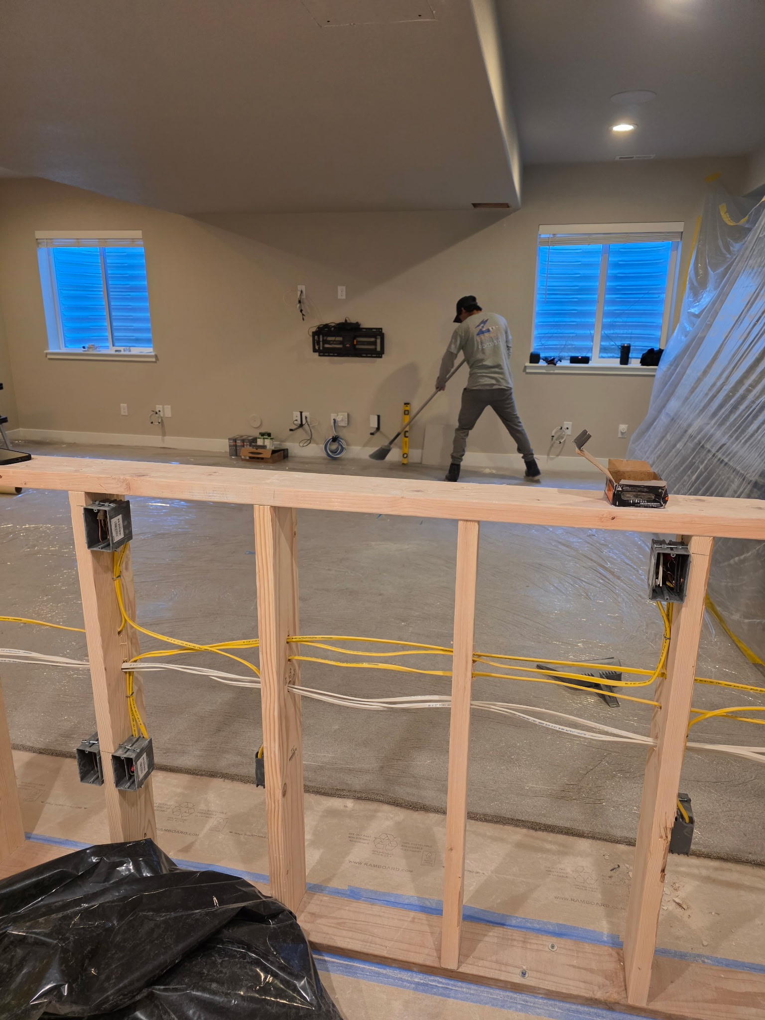 A construction worker smooths concrete flooring in a partially finished room with exposed framing and electrical wiring in the foreground.