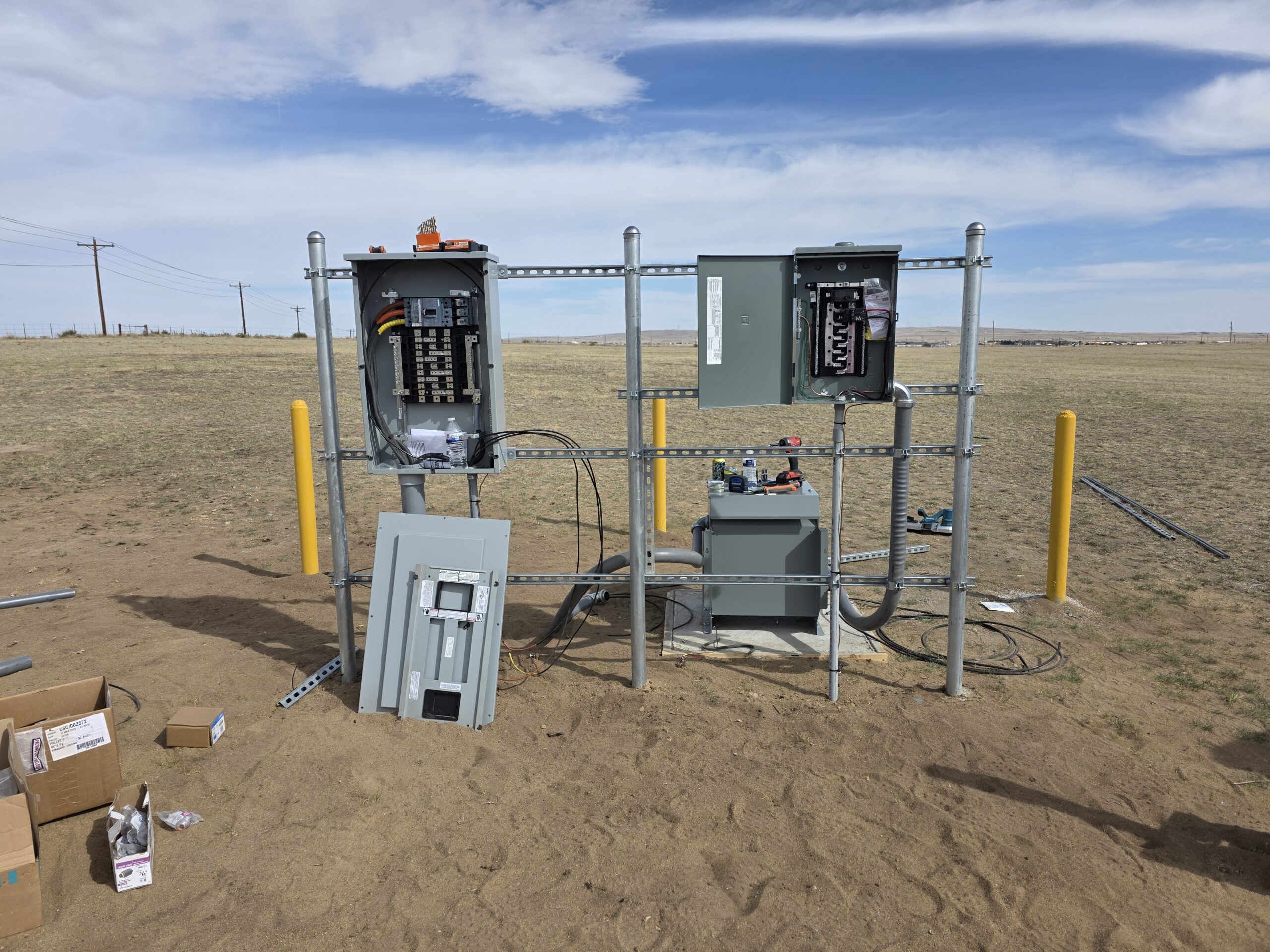 Outdoor electrical panel installation in progress on a metal frame in a sandy open field, with some electrical boxes open and tools scattered on the ground.
