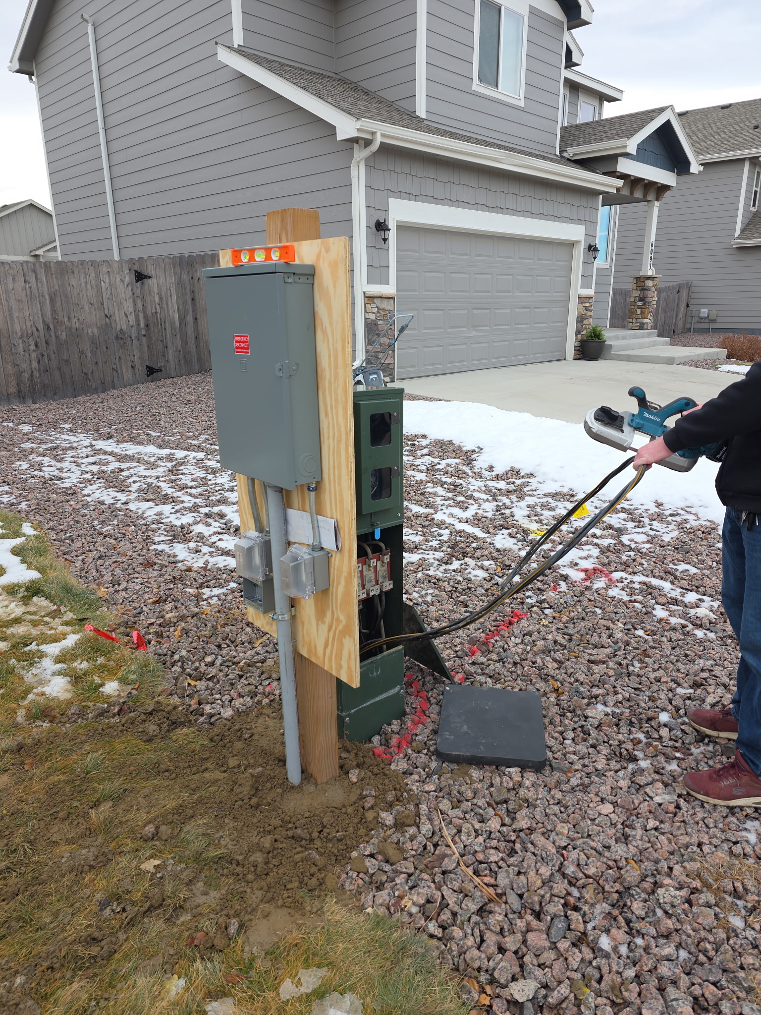 A person is holding cables near an open utility box and electrical panel on a residential lawn with some snow on the ground.