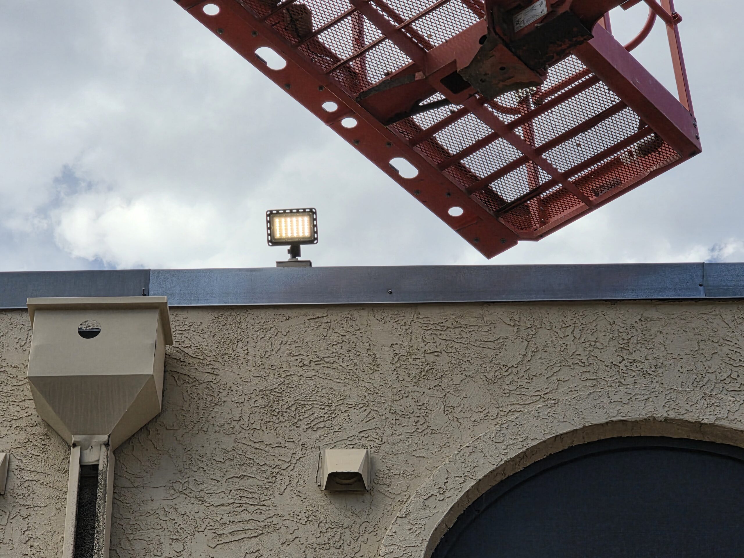 A red construction lift extends above a beige stucco building with exterior lights mounted near the roof against a cloudy sky.