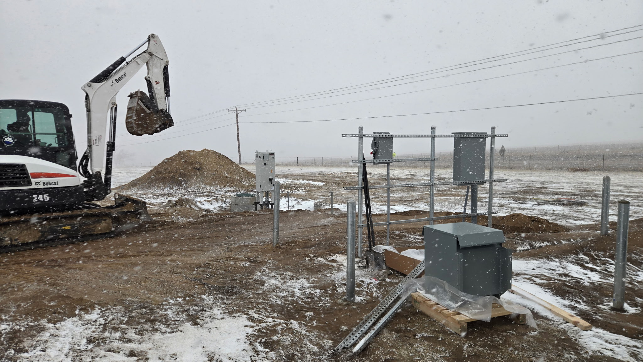 Small excavator moves dirt near electrical panels and utility boxes at a snowy outdoor construction site with metal posts and power lines visible in the background.
