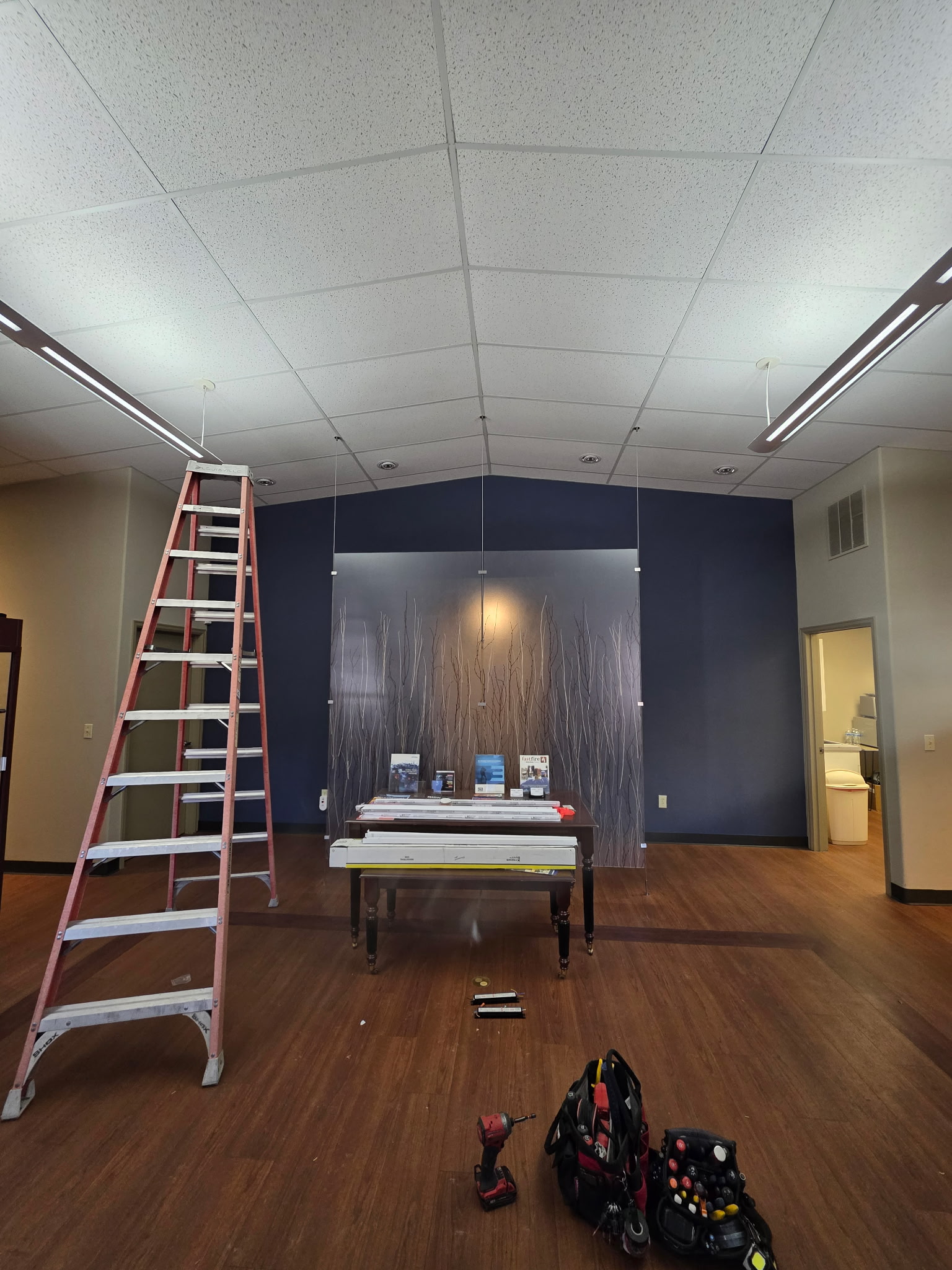 A red ladder stands next to a table with tools and equipment in a room with wood flooring and a blue accent wall featuring a decorative panel.