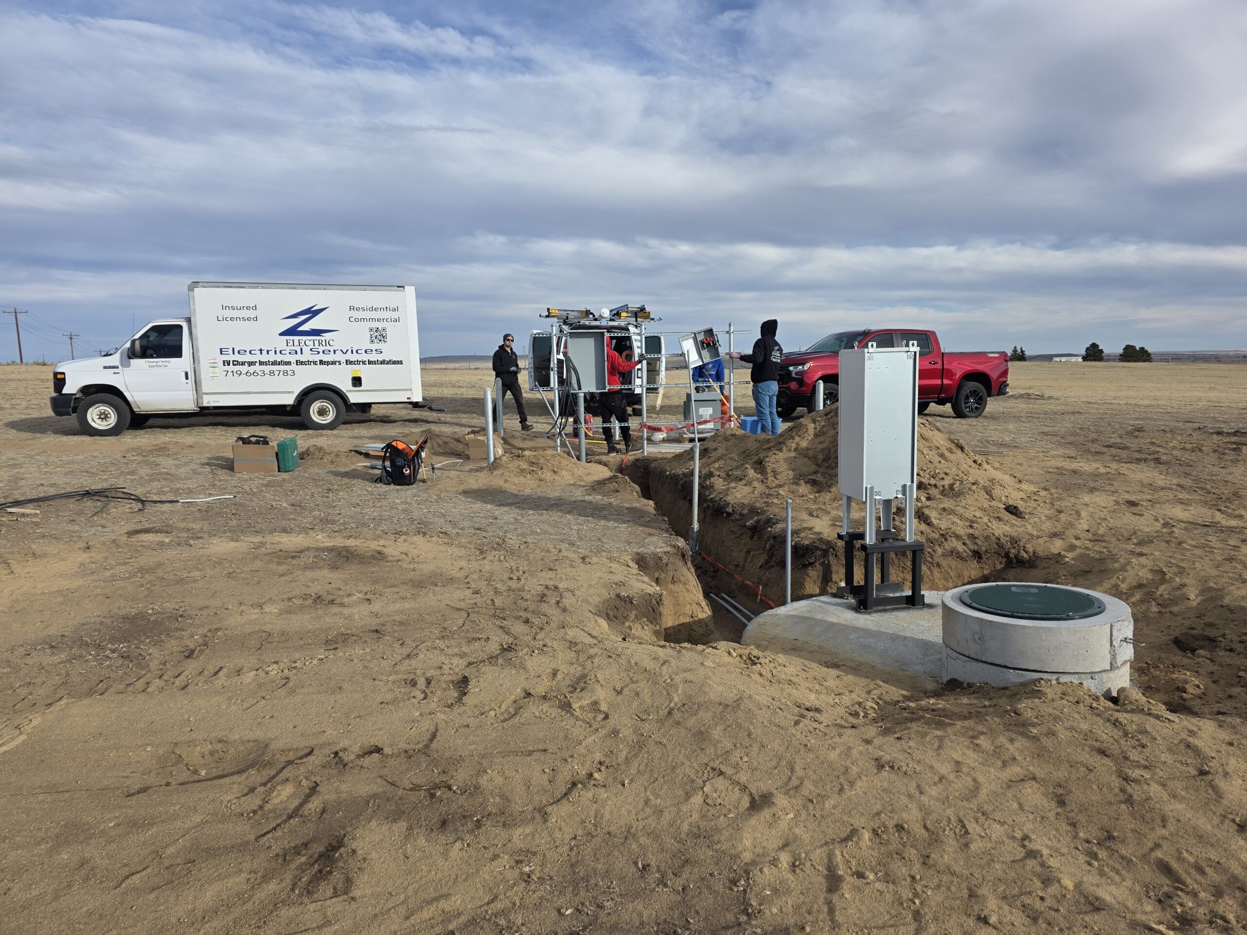 Workers stand near utility trucks at a construction site with an open trench, electrical equipment, and a utility box in a sandy outdoor area.