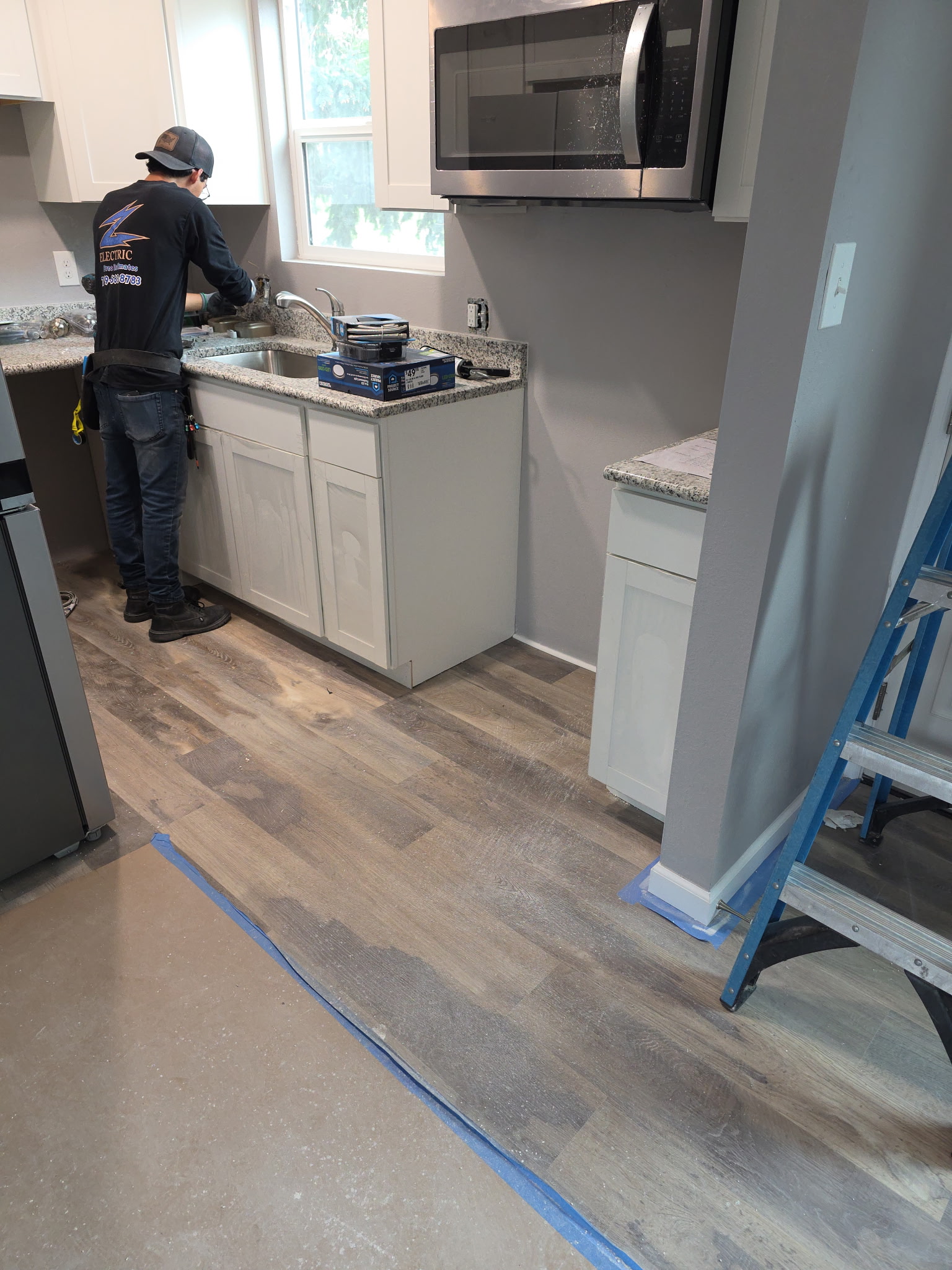 A worker installs or repairs a kitchen sink, with cabinets, granite countertops, and new flooring. A stepladder and masking tape are visible on the floor.