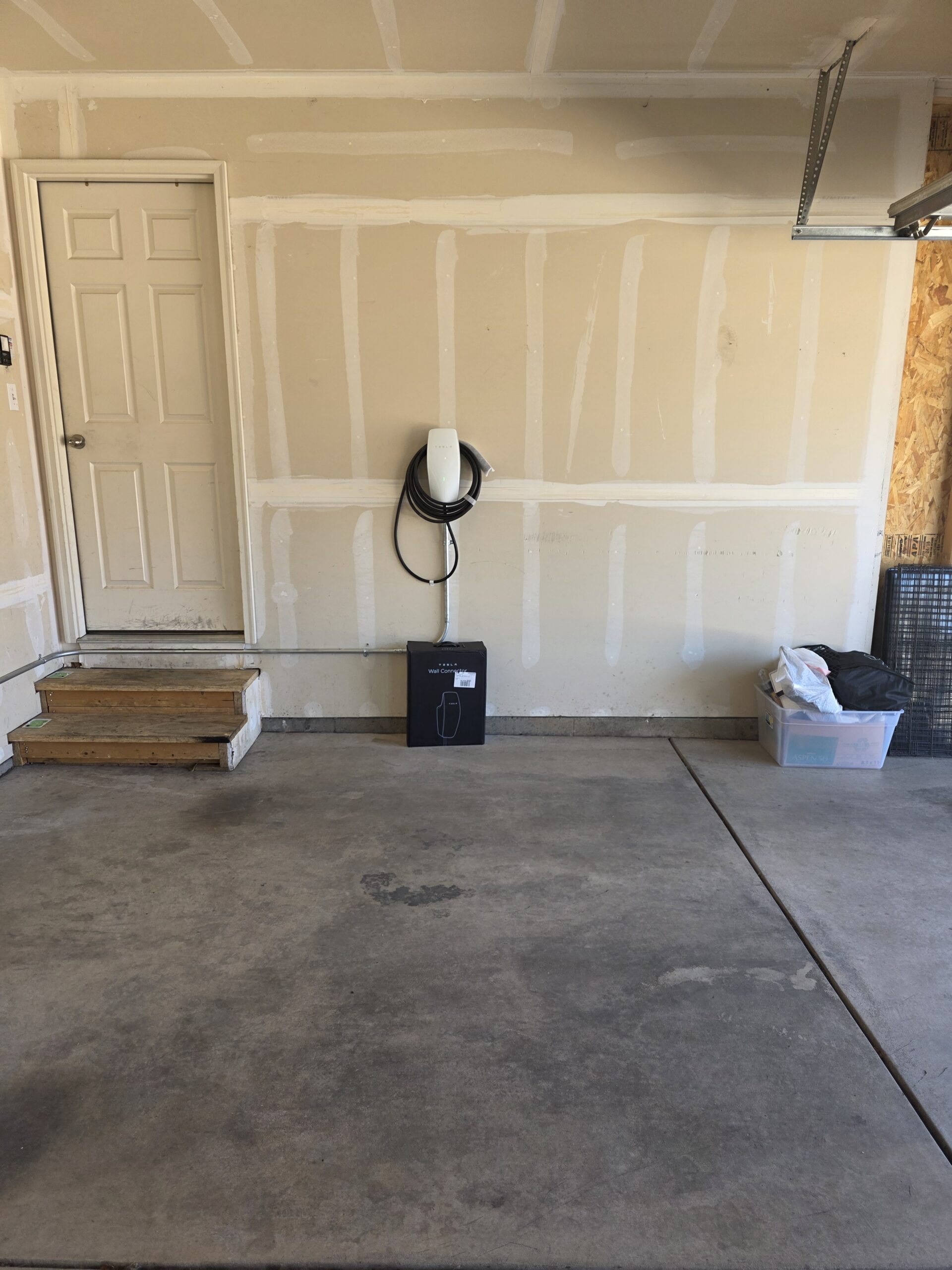 A garage with an electric vehicle charging station mounted on the wall, a box beneath it, steps leading to a door, and a plastic storage bin on the right.