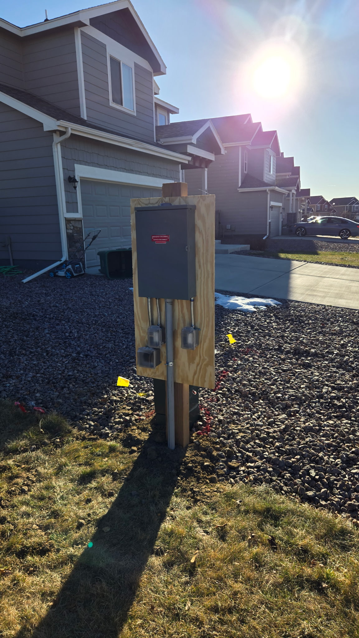 An electrical utility box mounted on wooden posts stands in front of a residential house on a sunny day.