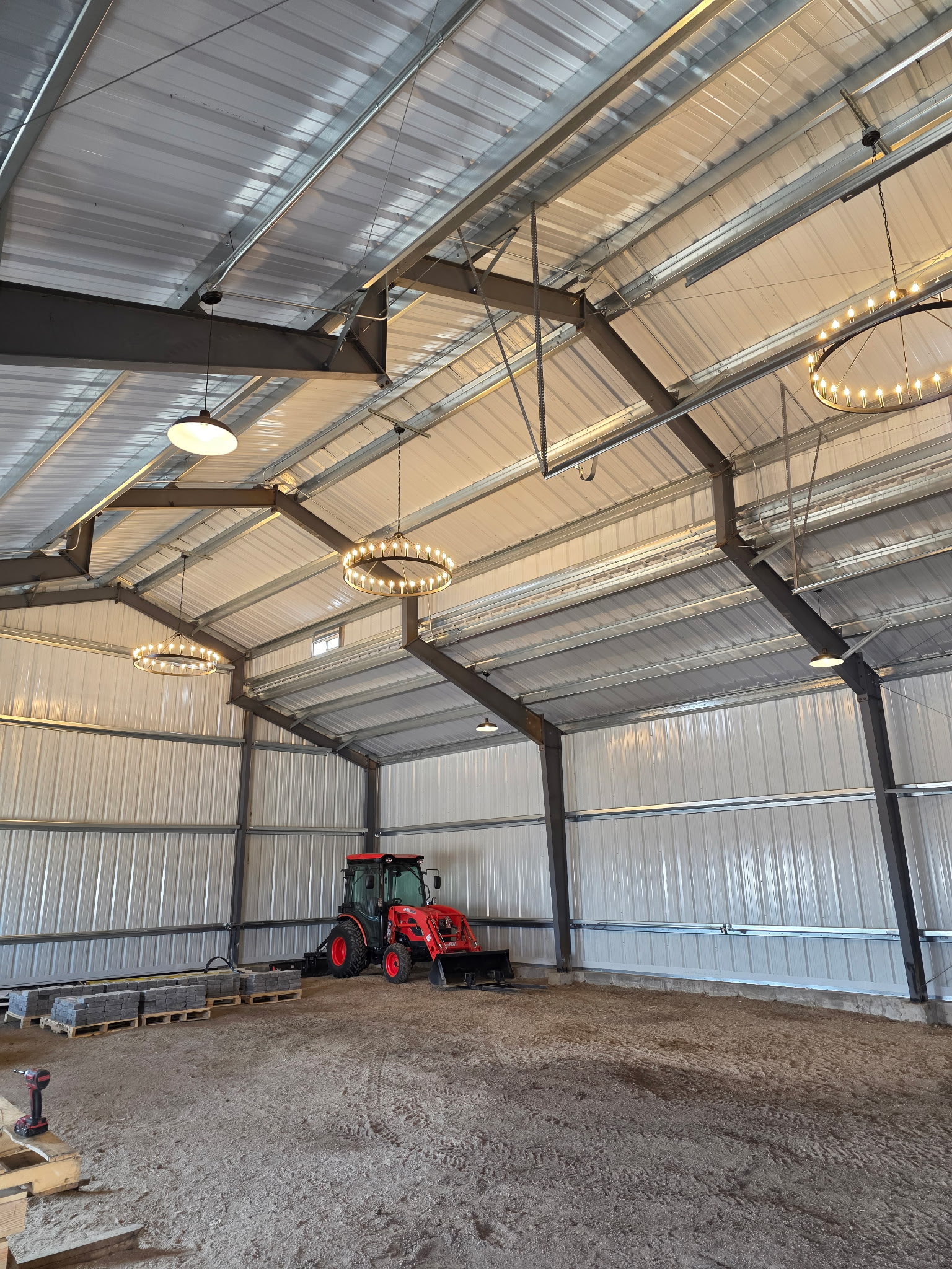 A red tractor is parked on a dirt floor inside a large metal barn with high ceilings and industrial light fixtures.