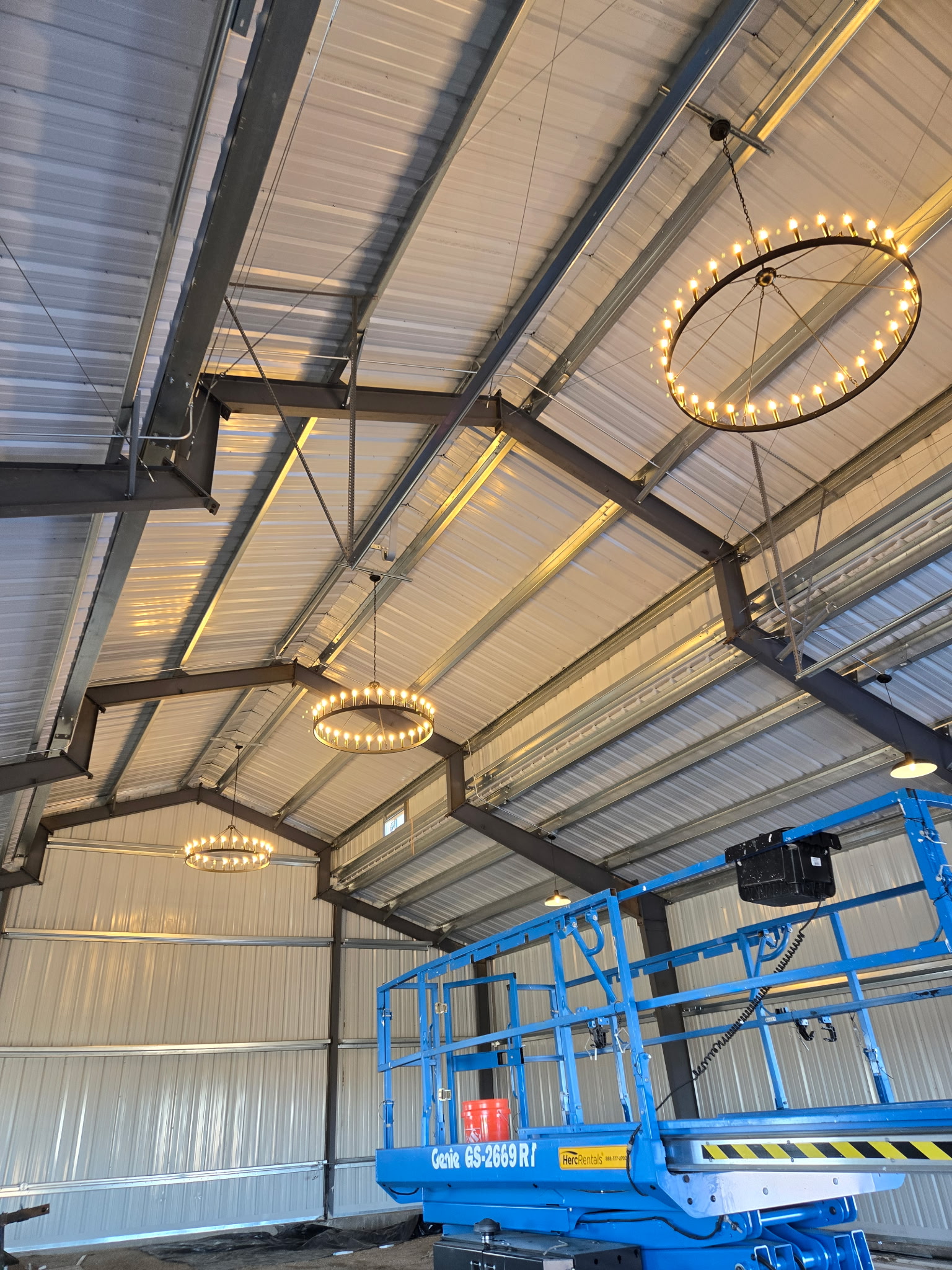 Interior of a metal building with exposed beams, three circular chandeliers with small bulbs, and a blue scissor lift parked inside.
