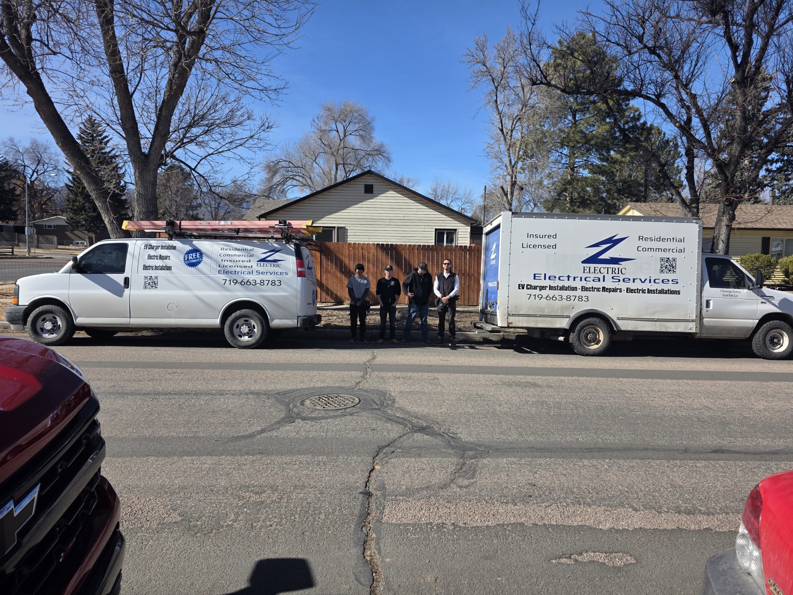 Two work vans with Electric Relief Services logos are parked on a residential street; four people stand between the vans in front of a house and wooden fence.