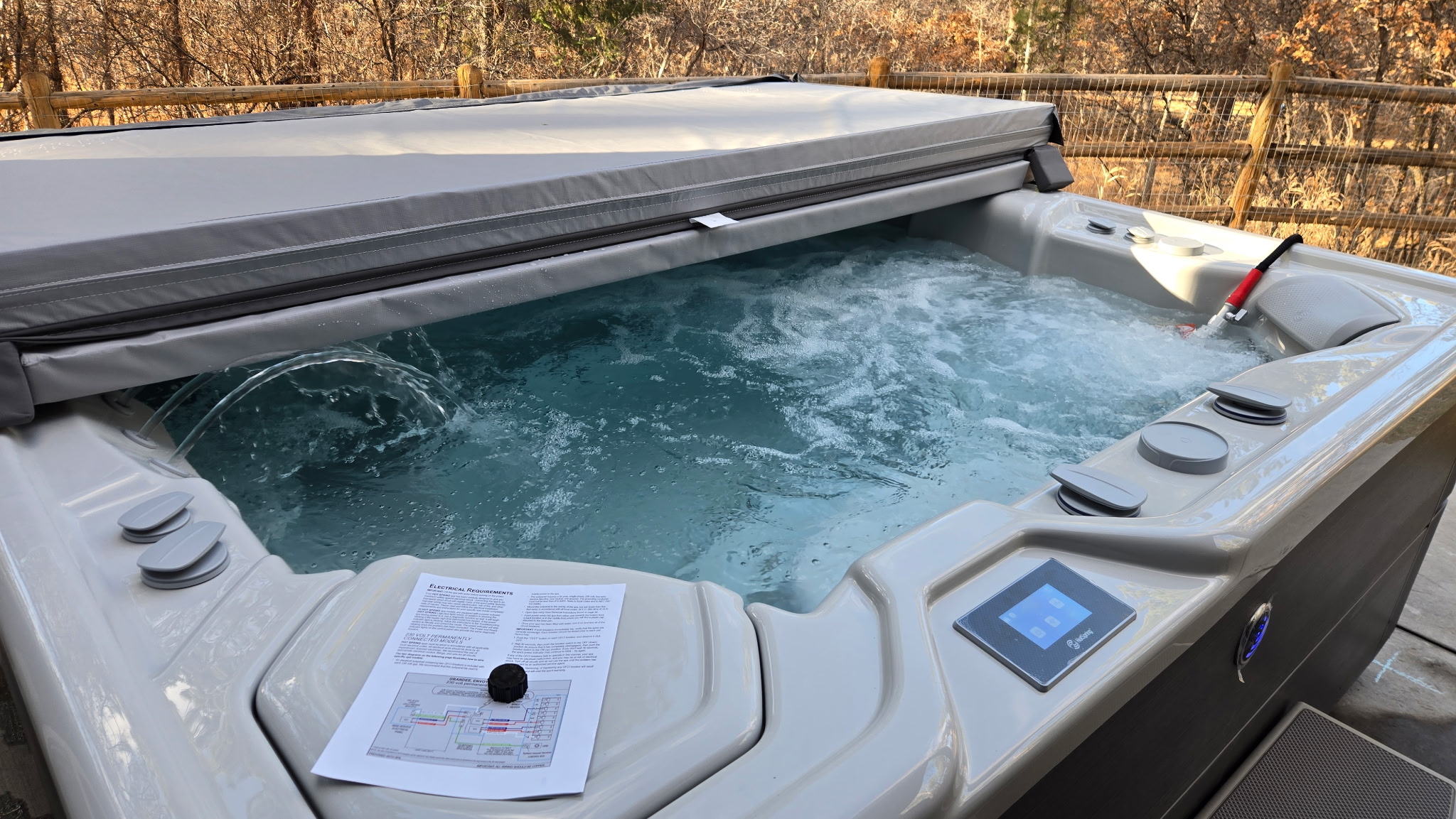 A bubbling outdoor hot tub with a partially closed cover, an instruction manual, Hot Tub Electrical Hookup details, a knob on the corner, and a control panel on the side. Trees and a wooden fence are visible in the background.