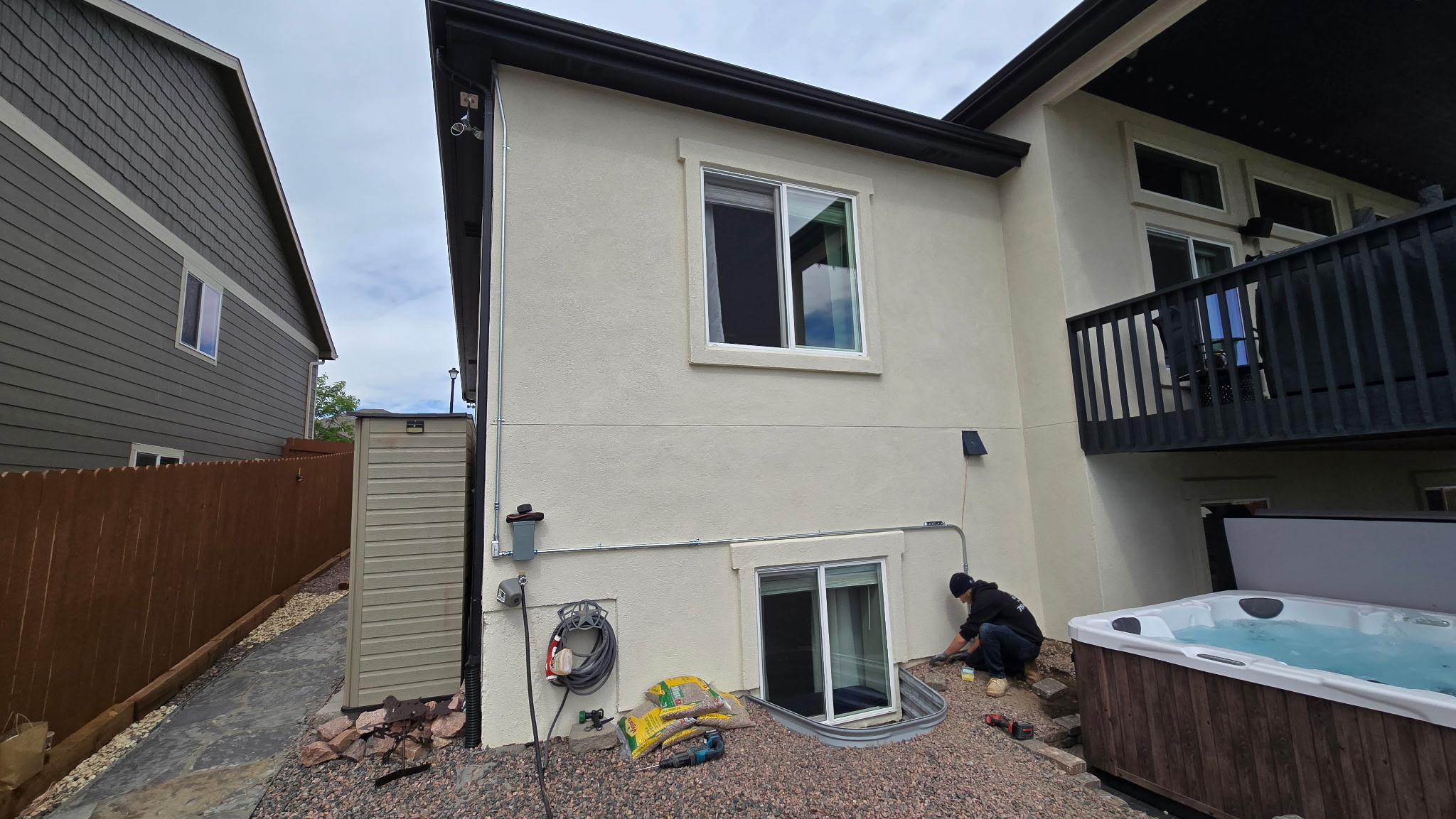 A person is working next to a basement window on the side of a two-story house with white stucco walls, possibly preparing for a hot tub electrical hookup; a hot tub and gardening items are nearby.