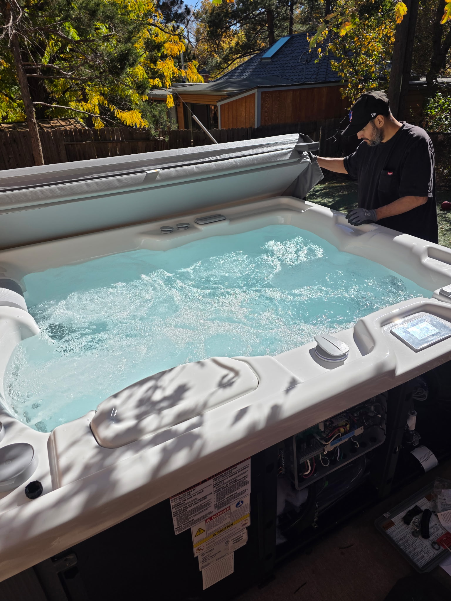 A person in a cap lifts the cover of a bubbling outdoor hot tub in a backyard on a sunny day, enjoying the relaxing benefits that hot tubs and spas offer.