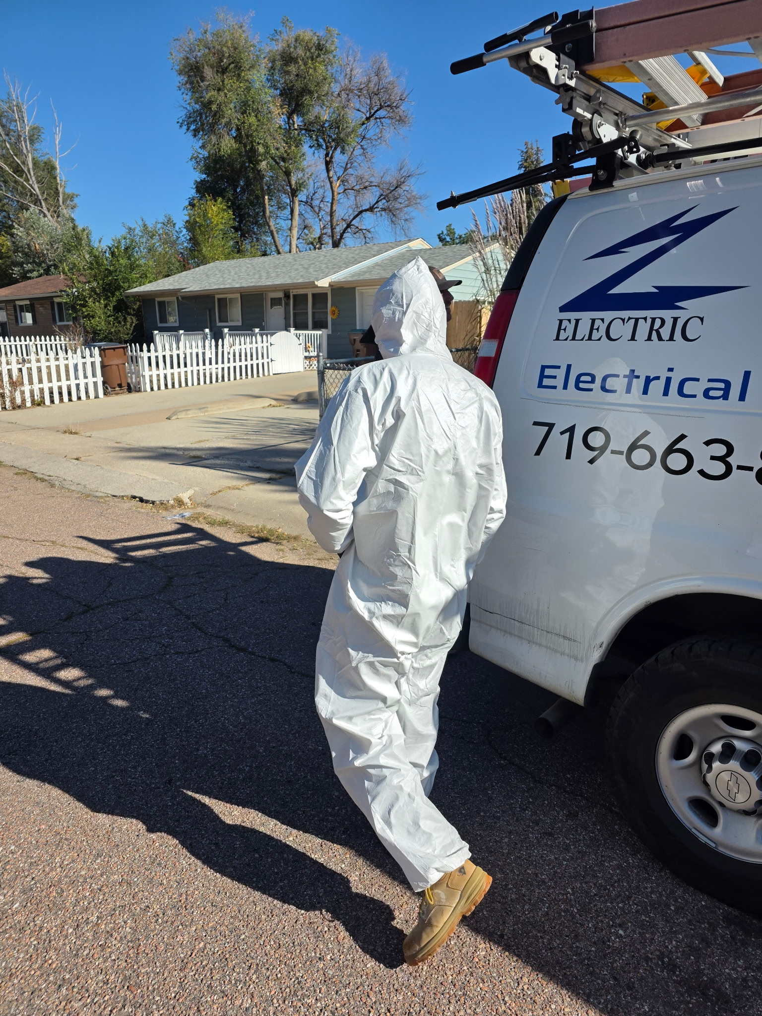Person in a white protective suit stands next to an electrical service van on a residential street, with houses and trees in the background.