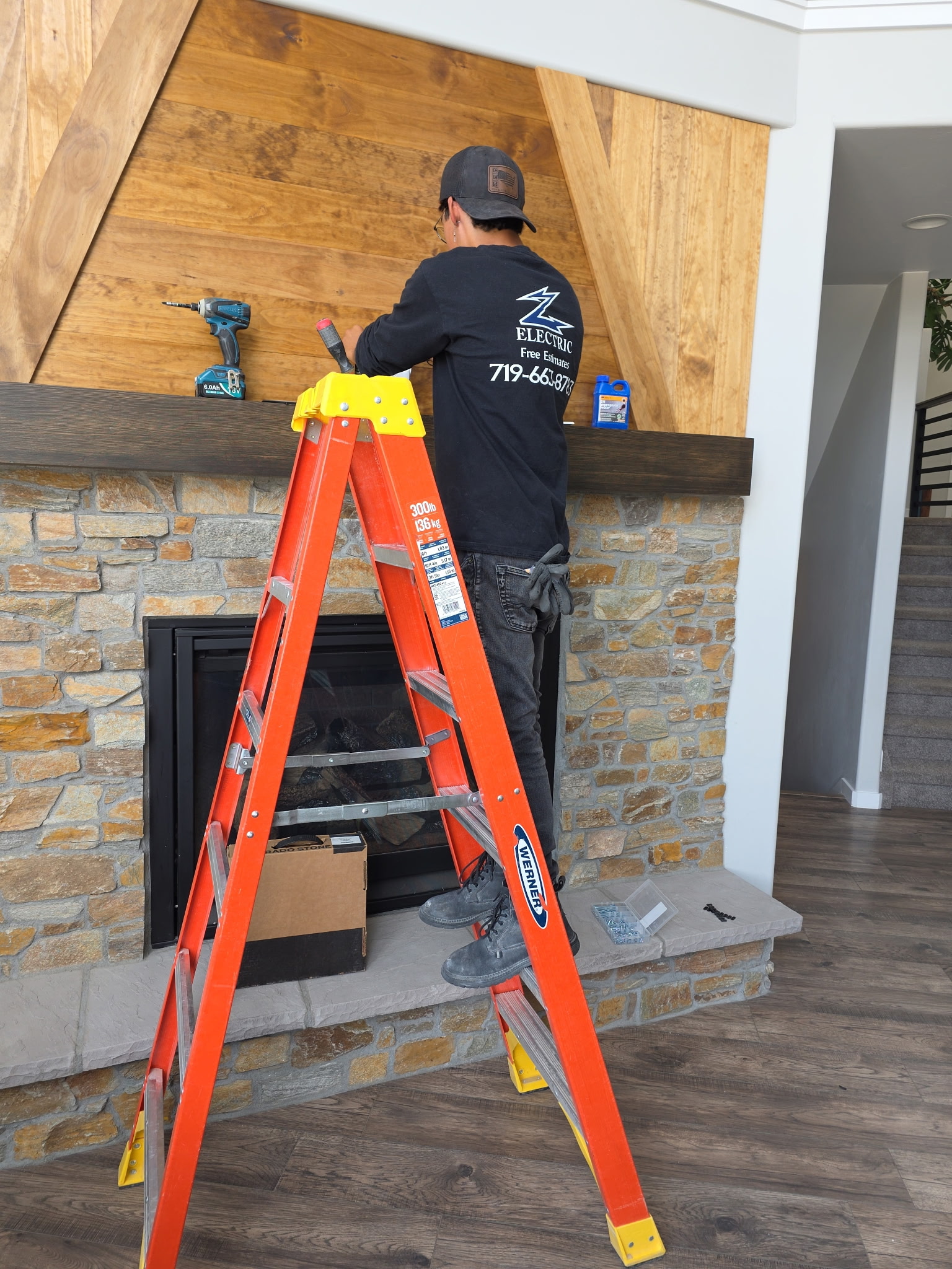 Person standing on a step ladder installs or repairs wood paneling above a fireplace, with tools and a toolbox nearby.