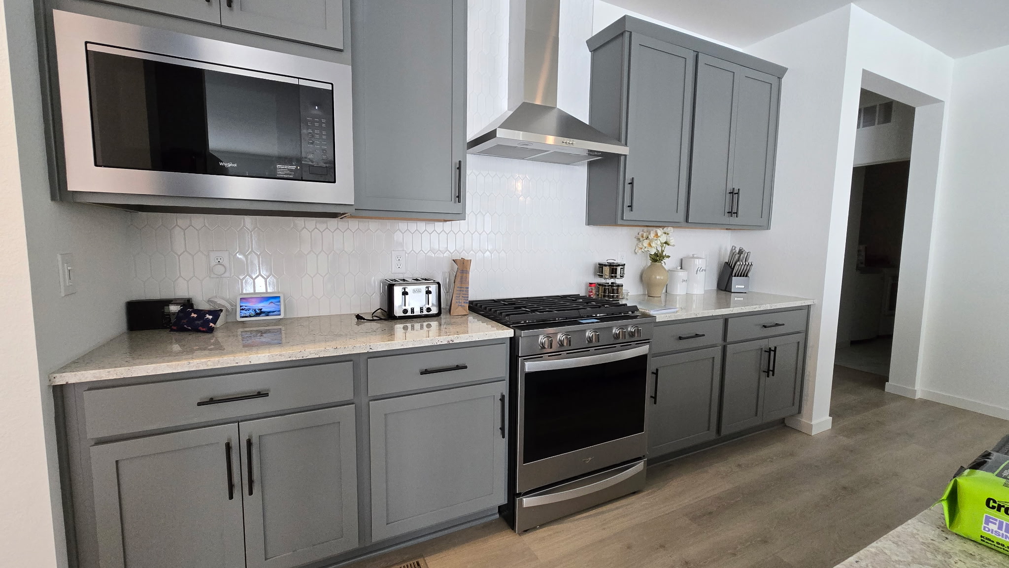 Modern kitchen with gray cabinets, stainless steel microwave and stove, white backsplash, countertop appliances, and decorative items on a light wood floor.