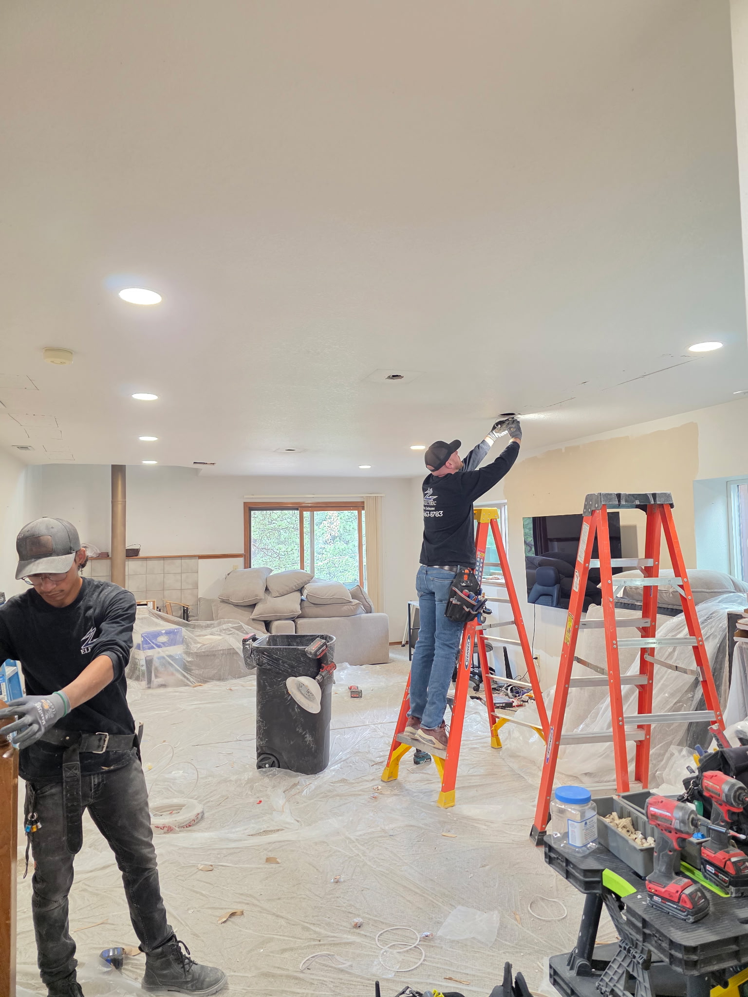 Two workers renovate a living room, one standing on a ladder installing a ceiling fixture, the other preparing materials; furniture is covered with plastic sheets.