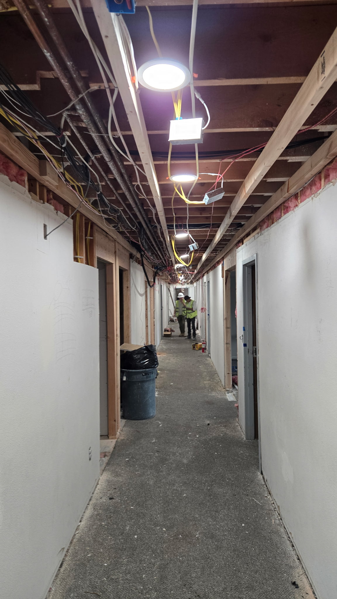 A hallway under construction with exposed ceiling wiring, temporary lighting, unfinished walls, debris on the floor, and a worker in safety gear standing at the far end.