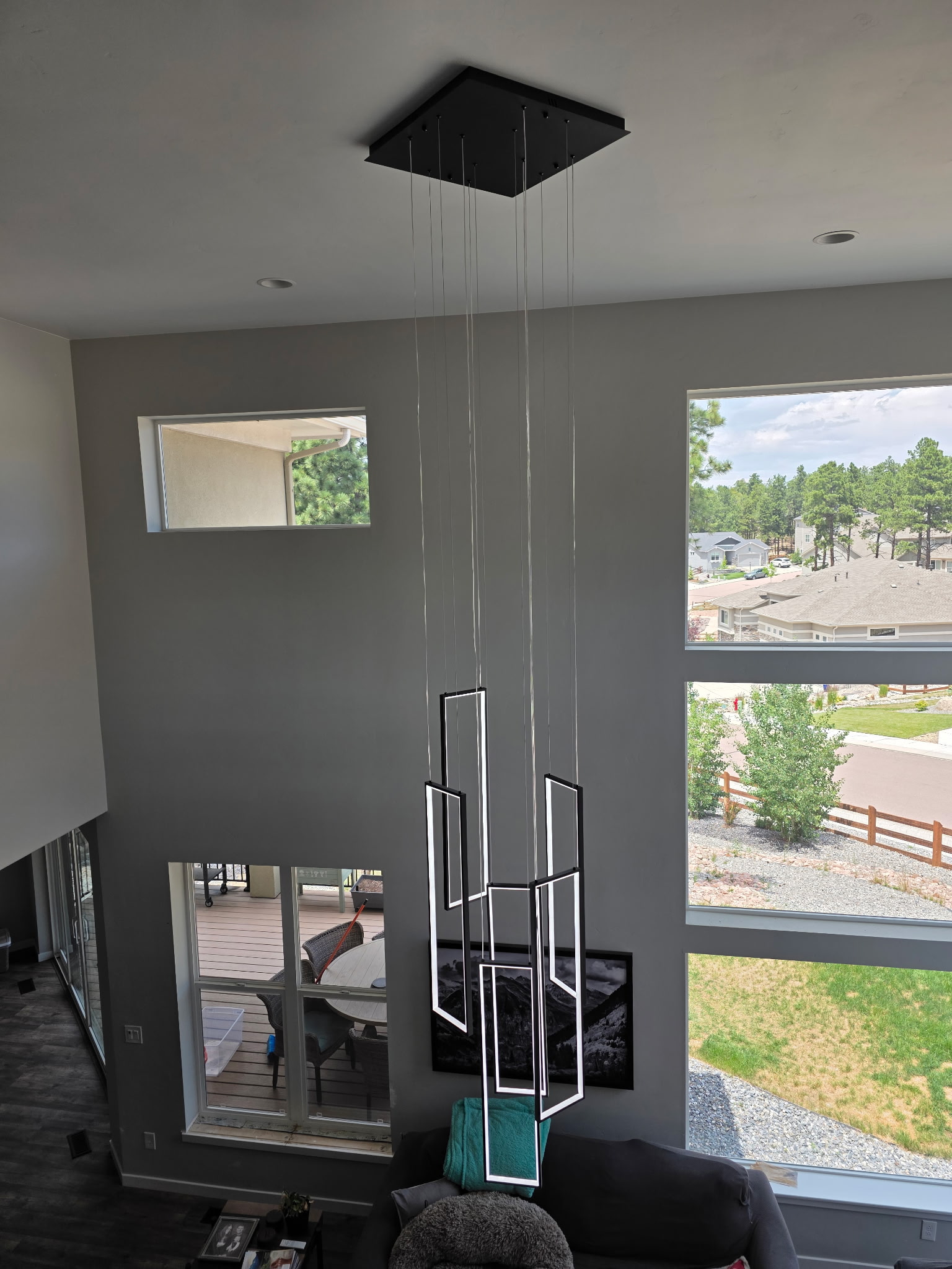 Modern geometric pendant chandelier with rectangular LED frames hanging from a high ceiling in a bright living room with large windows and outdoor views.