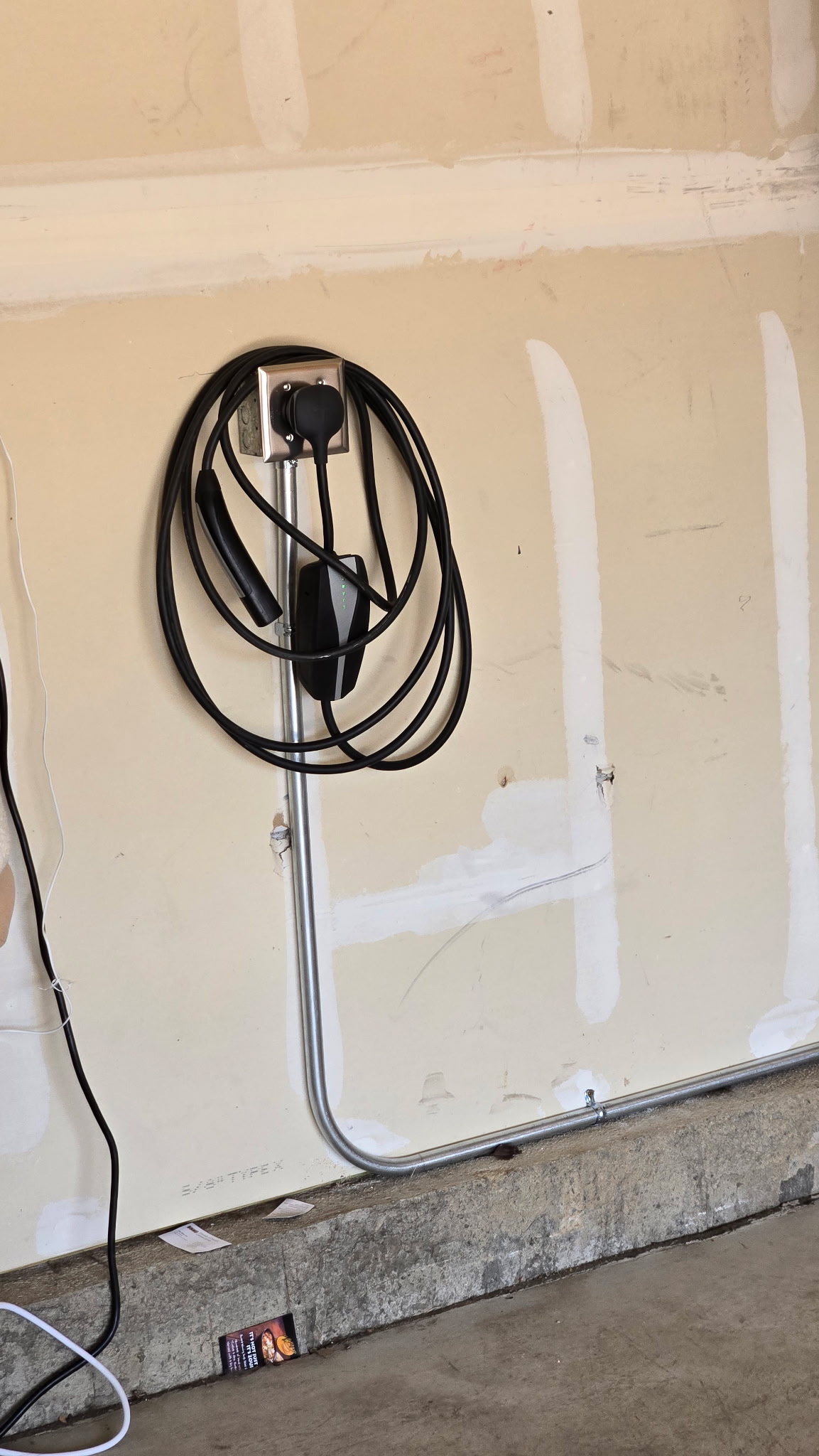A black electric vehicle charging cable is coiled on a wall-mounted charger in a garage with unfinished drywall.