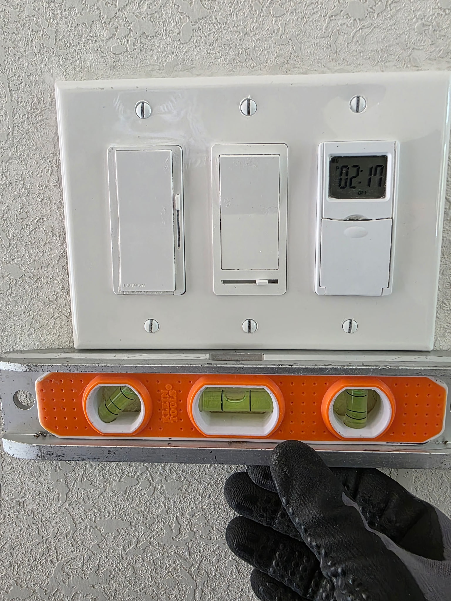 A gloved hand holds a level under a light switch panel with three switches and a digital timer on a textured wall; the level shows the panel is not horizontal.