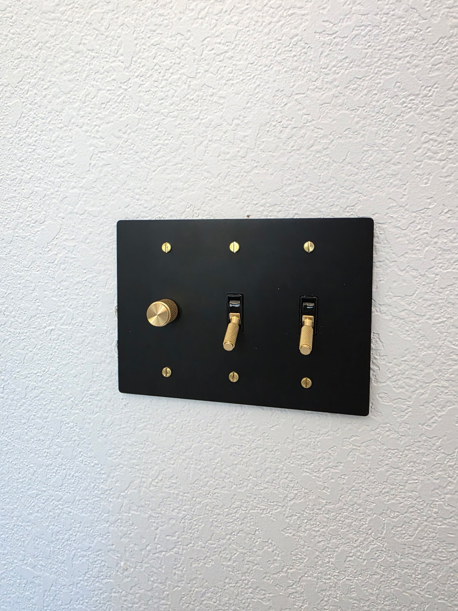 A black switch plate with two brass toggle switches and one brass rotary dimmer is mounted on a textured white wall.