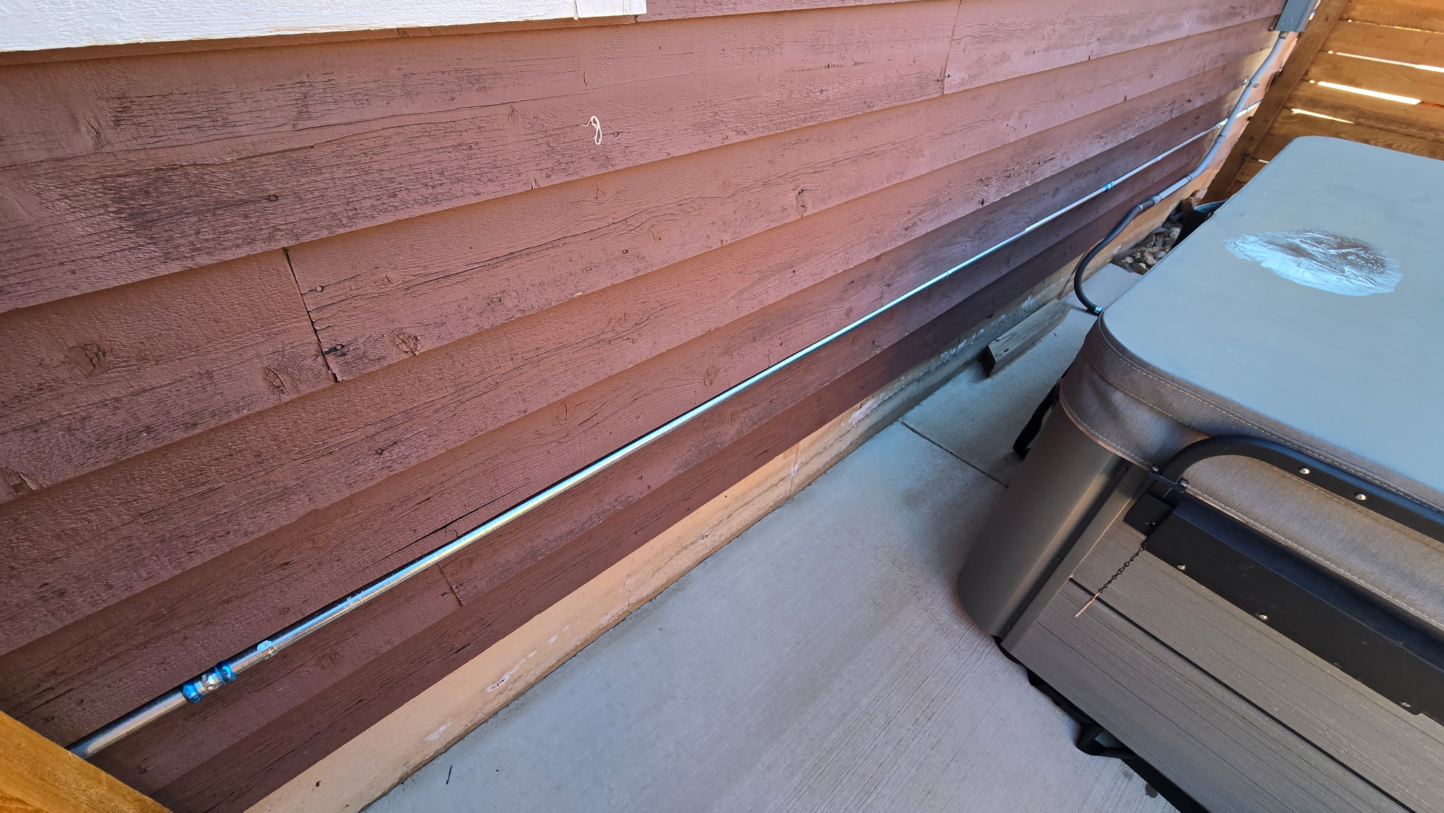 A metal conduit runs horizontally along a wooden exterior wall next to a hot tub on a concrete patio.