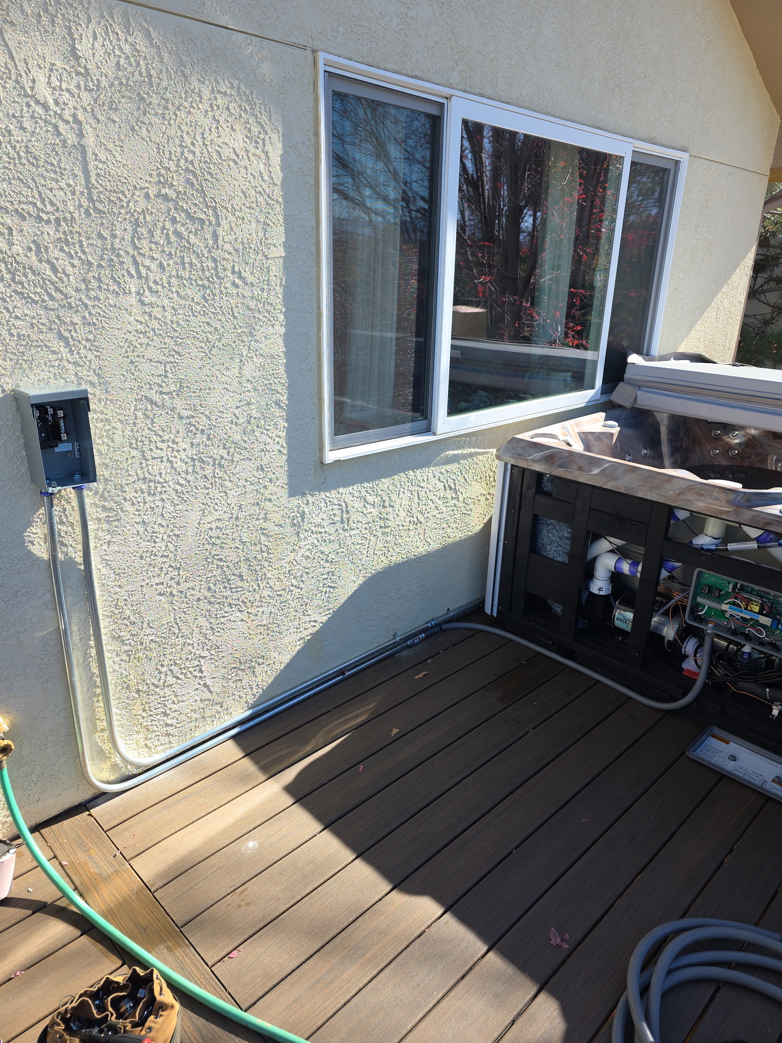 A hot tub with its side panel open is on a wooden deck next to a beige stucco wall with a window and an outdoor electrical box. A garden hose is on the deck.
