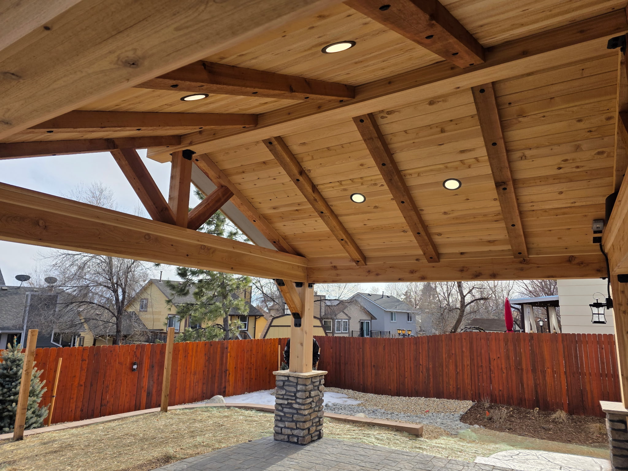 A wooden patio cover with exposed beams and recessed lights stands over a paved area in a backyard with a red wooden fence and neighboring houses visible.