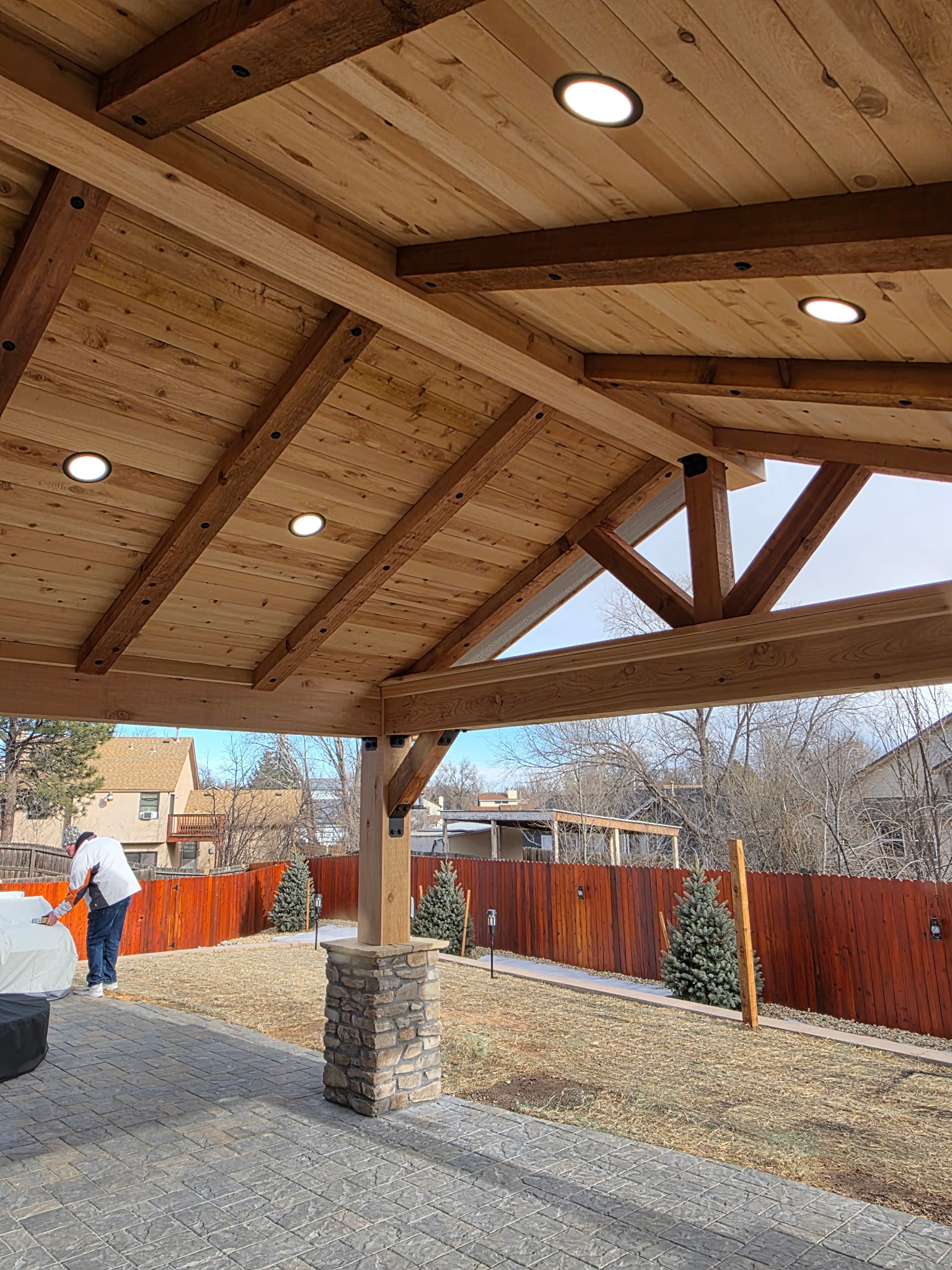 A wooden patio cover with exposed beams, stone pillars, and recessed lights overlooks a fenced backyard with sparse grass and small trees; a person stands near covered furniture.