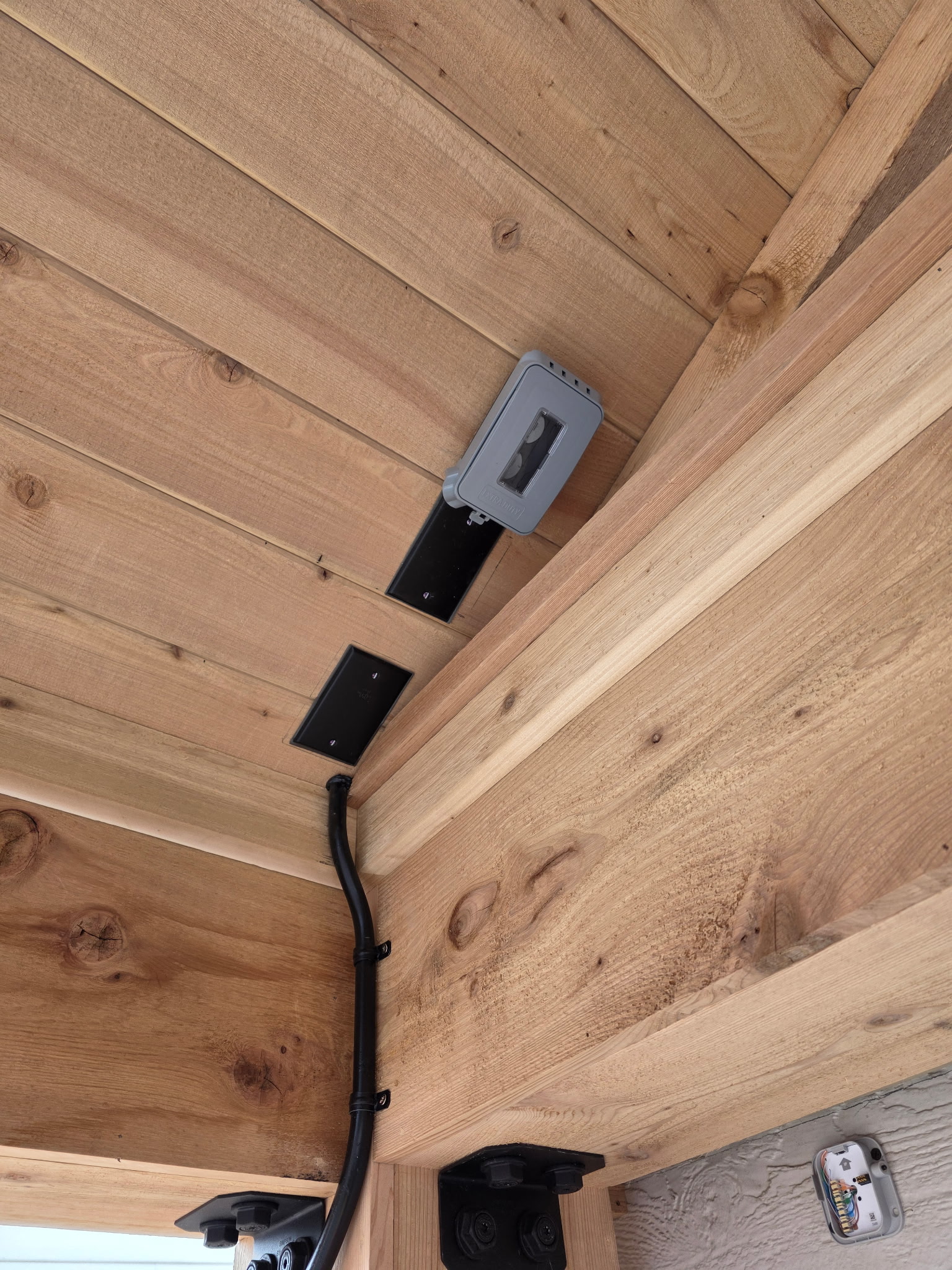 Ceiling corner with exposed wood panels, electrical conduit, two black electrical boxes, and a gray weatherproof outlet cover.