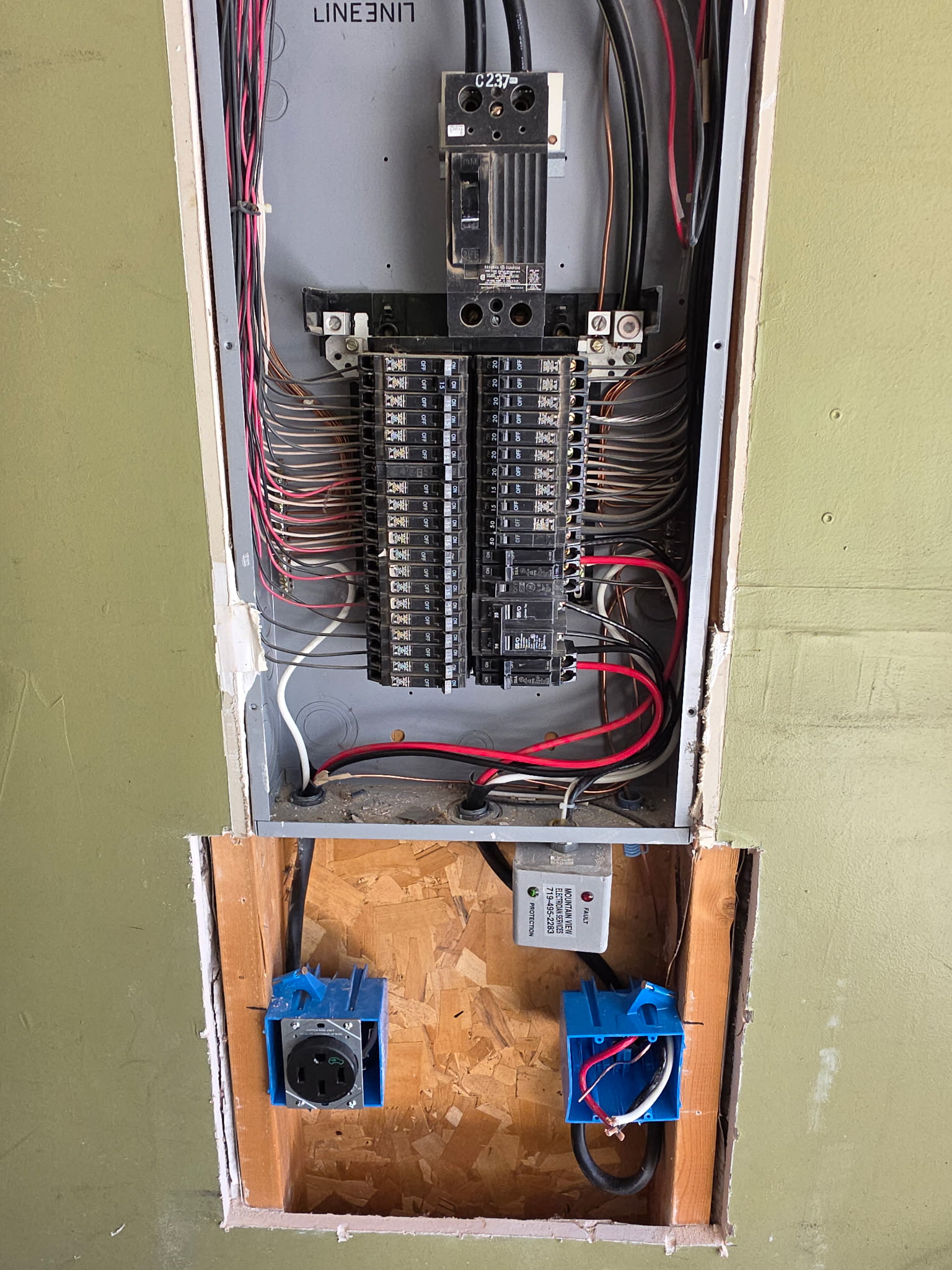 An open electrical panel with exposed wiring and circuit breakers, and two blue electrical outlet boxes in a cutout below the panel.