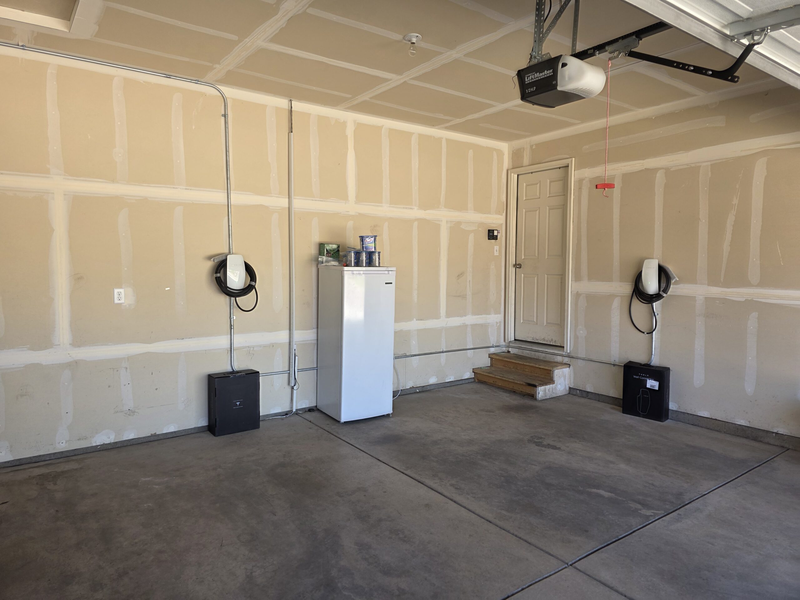 A garage with unfinished walls, a white refrigerator, two Tesla Wall Connector Installation units mounted on the walls, steps leading to a door, and a partially open garage door.