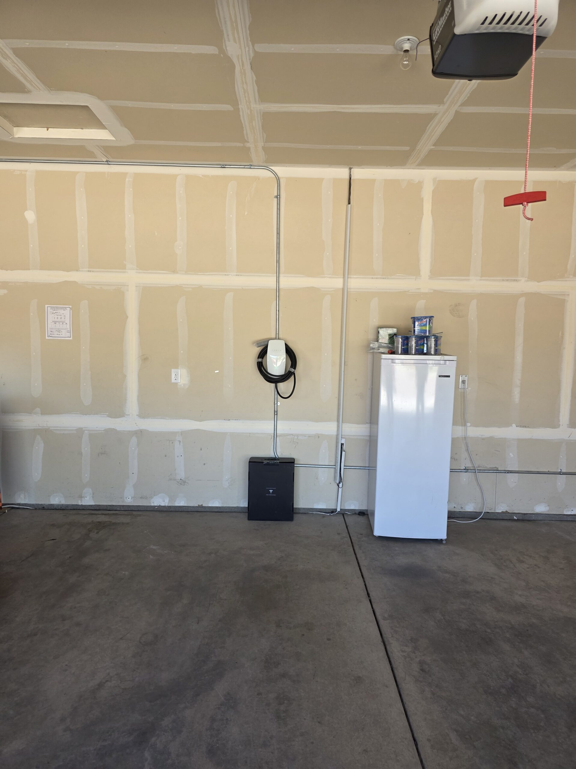 A white refrigerator and several cans are positioned against an unfinished garage wall with an electric car charger mounted beside them.