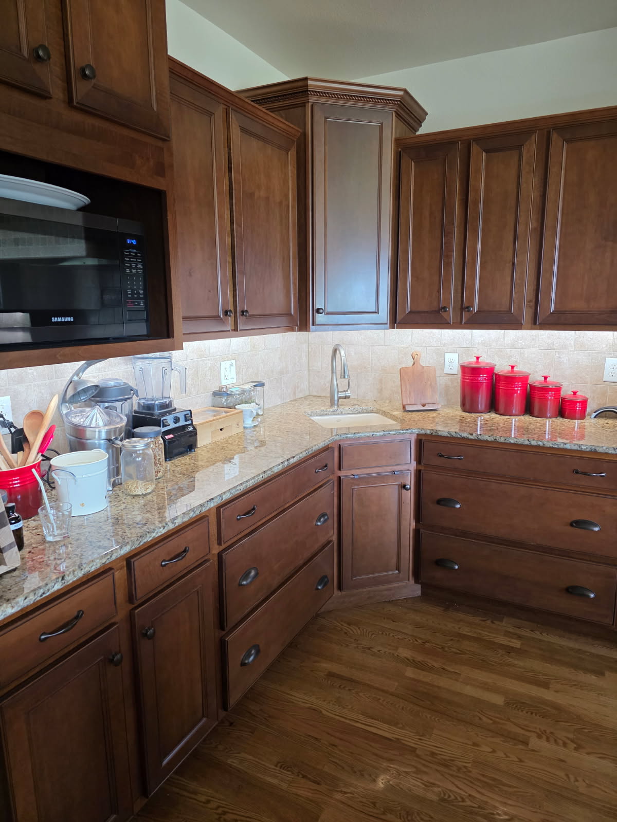 A kitchen with wooden cabinets, granite countertops, a microwave, blender, toaster, red storage canisters, and various kitchen items on the counters features under cabinet lighting installation for added brightness.
