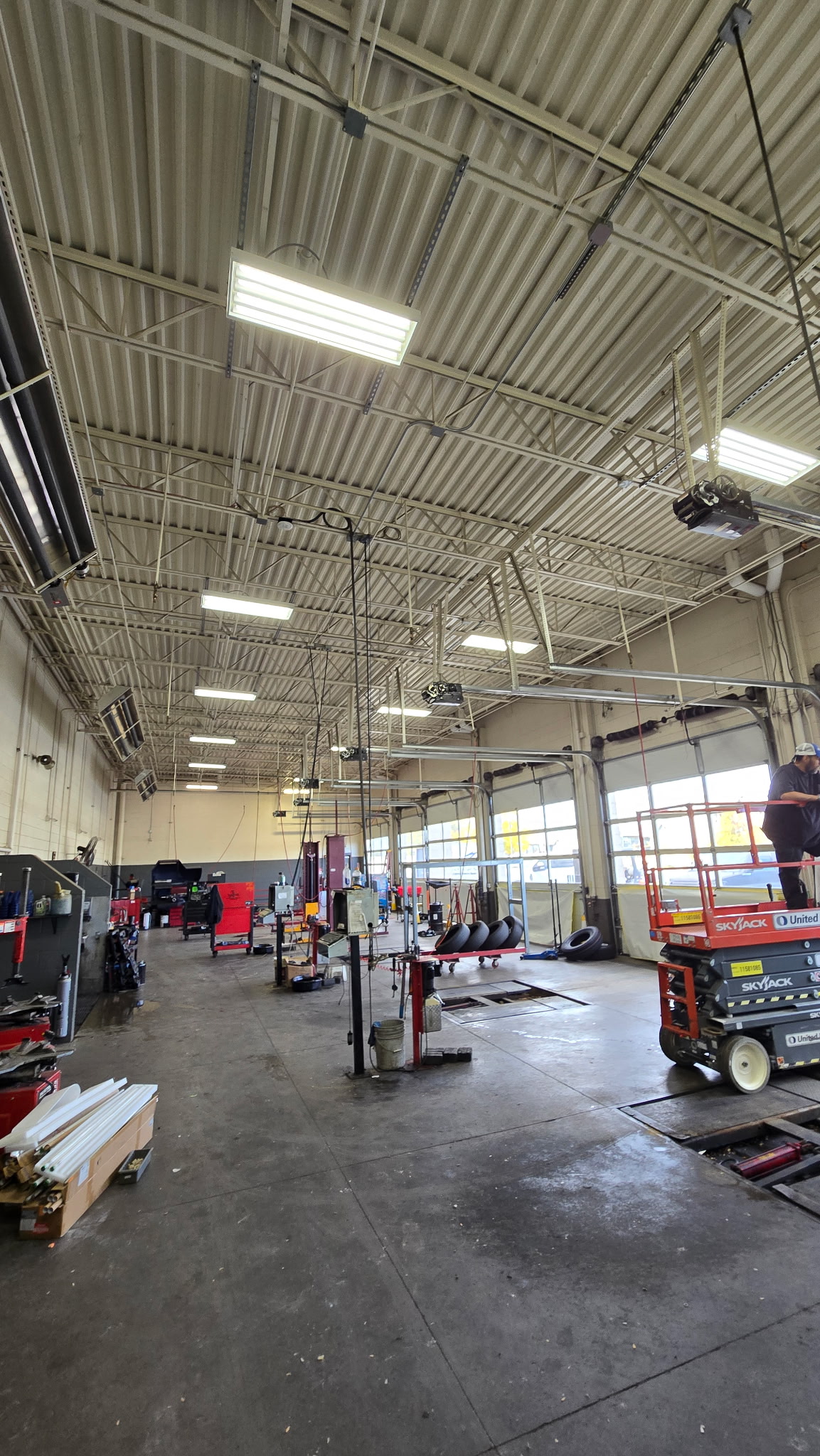A spacious auto repair shop interior with high ceilings, equipment, tools, and machinery; a worker stands on a lift at the right side of the image.