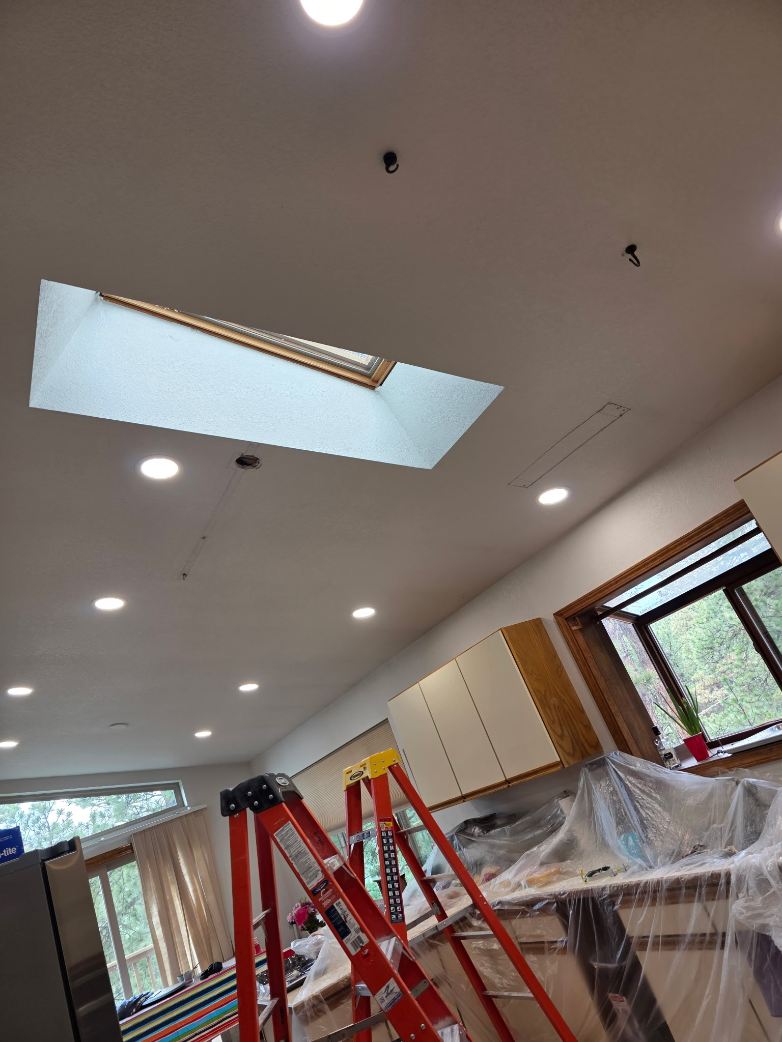 A kitchen under renovation with covered cabinets, a skylight, several ceiling lights, and two red ladders.